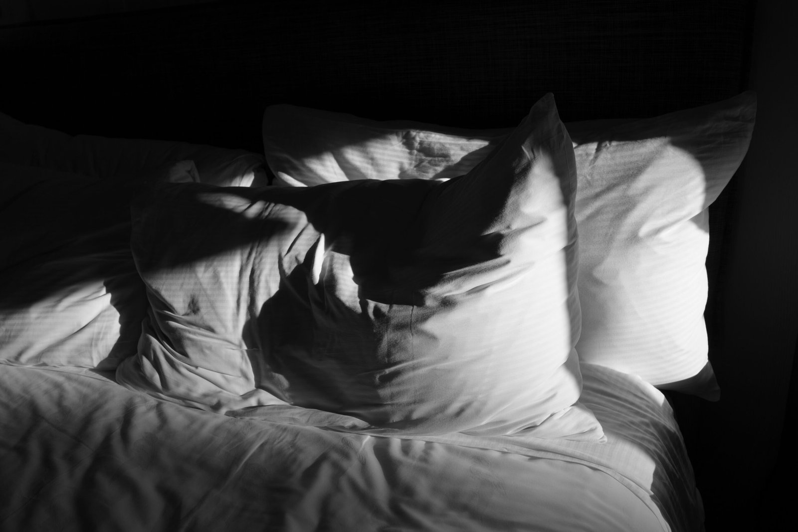 Soft white pillows on a bed with morning light, representing the need for darkness tools like sleep masks or blackout curtains
